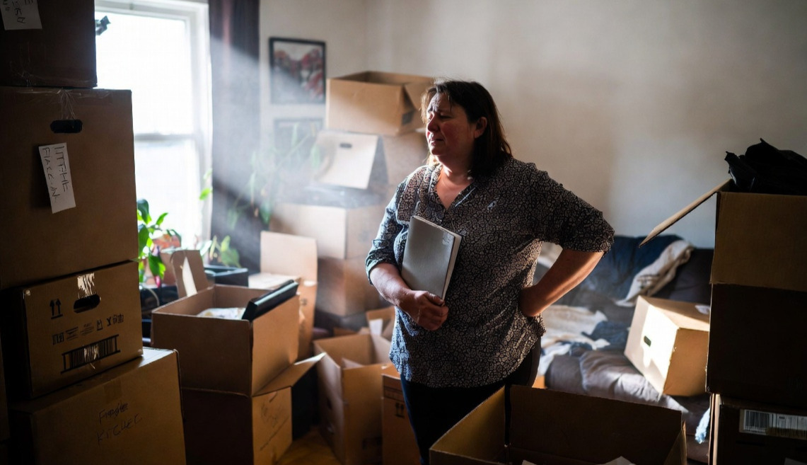A woman stands in a cluttered living room full of moving boxers with a look of overwhelmed grief on her face.